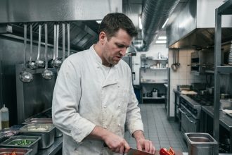 Chef cuisinier professionnel en veste blanche découpant des légumes dans une cuisine de restaurant avec plan de travail en inox