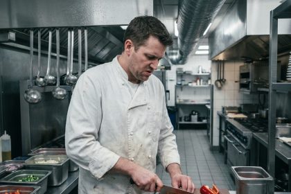 Chef cuisinier professionnel en veste blanche découpant des légumes dans une cuisine de restaurant avec plan de travail en inox